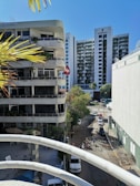A cityscape featuring a street lined with modern buildings, including a high-rise named Emporio. Several vehicles are parked along the road and pedestrians are visible. The foreground shows part of a balcony railing and palm leaves, indicating a tropical or warm climate.