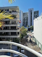 A cityscape featuring a street lined with modern buildings, including a high-rise named Emporio. Several vehicles are parked along the road and pedestrians are visible. The foreground shows part of a balcony railing and palm leaves, indicating a tropical or warm climate.