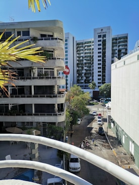 A cityscape featuring a street lined with modern buildings, including a high-rise named Emporio. Several vehicles are parked along the road and pedestrians are visible. The foreground shows part of a balcony railing and palm leaves, indicating a tropical or warm climate.