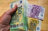 A hand is holding a large stack of 100 euro banknotes with additional 500 and 200 euro notes placed on a wooden surface in the background.