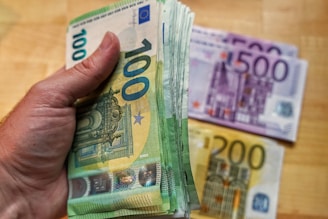 A hand is holding a large stack of 100 euro banknotes with additional 500 and 200 euro notes placed on a wooden surface in the background.