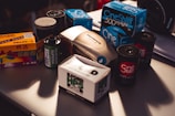 A collection of vintage film photography equipment including various boxes and rolls of film, an Olympus film camera, and film canisters. The brands visible include Kodak, Fujicolor, Ilford, and CineStill, and they are placed on a table with subdued lighting.
