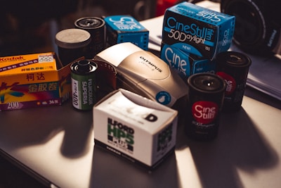A collection of vintage film photography equipment including various boxes and rolls of film, an Olympus film camera, and film canisters. The brands visible include Kodak, Fujicolor, Ilford, and CineStill, and they are placed on a table with subdued lighting.