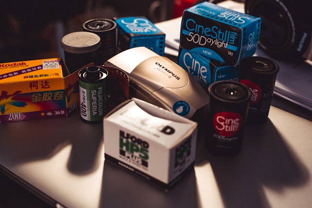 A collection of vintage film photography equipment including various boxes and rolls of film, an Olympus film camera, and film canisters. The brands visible include Kodak, Fujicolor, Ilford, and CineStill, and they are placed on a table with subdued lighting.