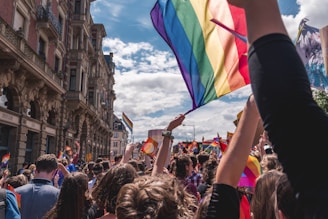 A vibrant crowd gathered for a parade or march on a city street. Many participants are holding rainbow flags representing LGBTQ+ pride. The buildings lining the street have ornate architectural details, and the sky is partly cloudy with a clear view overhead.