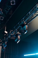 A series of stage lights are attached to a truss structure, with visible cables and metal components. The lights are positioned at various angles, suggesting a setup for a performance or event. The background includes darkened segments, emphasizing the focus on the lighting equipment.