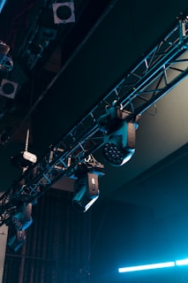 A series of stage lights are attached to a truss structure, with visible cables and metal components. The lights are positioned at various angles, suggesting a setup for a performance or event. The background includes darkened segments, emphasizing the focus on the lighting equipment.