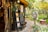 A quaint outdoor bookshop with colorful fabric drapes on the left, filled with books and decorative items. There's a blue ladder with chalkboards displaying written messages and various artwork. A headless statue wearing a suit stands in the garden area surrounded by greenery and trees with autumn foliage. A small pink round table sits nearby, adding to the whimsical atmosphere.