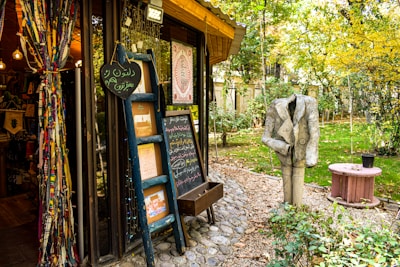 A quaint outdoor bookshop with colorful fabric drapes on the left, filled with books and decorative items. There's a blue ladder with chalkboards displaying written messages and various artwork. A headless statue wearing a suit stands in the garden area surrounded by greenery and trees with autumn foliage. A small pink round table sits nearby, adding to the whimsical atmosphere.