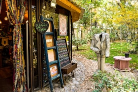 A quaint outdoor bookshop with colorful fabric drapes on the left, filled with books and decorative items. There's a blue ladder with chalkboards displaying written messages and various artwork. A headless statue wearing a suit stands in the garden area surrounded by greenery and trees with autumn foliage. A small pink round table sits nearby, adding to the whimsical atmosphere.