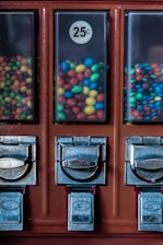 Three candy vending machine compartments filled with multicolored, round candy pieces, each priced at 25 cents. The machine features metal dispensers with a slightly worn appearance.
