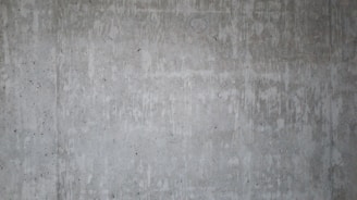 A textured concrete wall with visible streaks and patches of darker and lighter shades, showing natural imperfections and a raw industrial look.