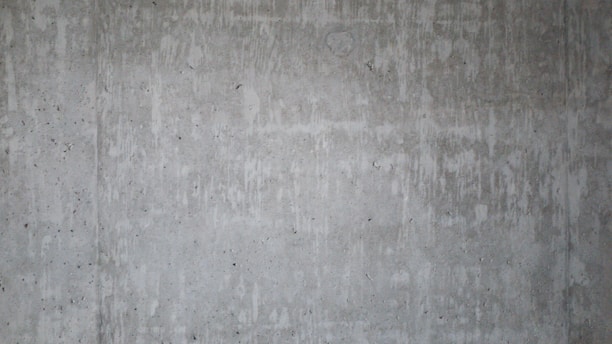 A textured concrete wall with visible streaks and patches of darker and lighter shades, showing natural imperfections and a raw industrial look.
