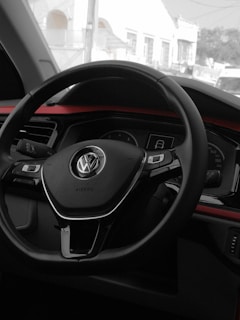 A close-up view of a car's interior, focusing on the steering wheel. The logo on the steering wheel indicates it belongs to Volkswagen. The dashboard and part of the front window are also visible, with the view outside showing some buildings.