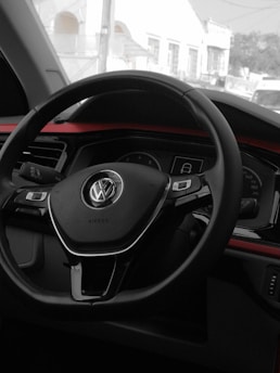 A close-up view of a car's interior, focusing on the steering wheel. The logo on the steering wheel indicates it belongs to Volkswagen. The dashboard and part of the front window are also visible, with the view outside showing some buildings.