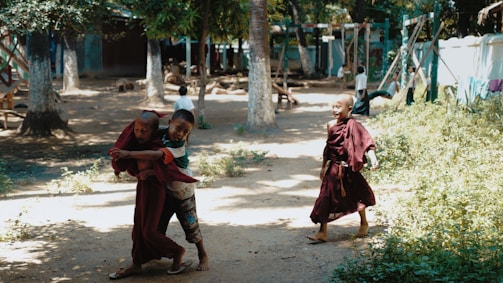 Children playing in an outdoor setting surrounded by trees and greenery. Some of them are wearing traditional monk robes and appear to be enjoying a sunny day, laughing and running.