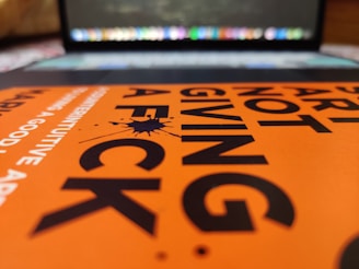 A close-up view of an orange book cover with bold black text, partially focusing on the words 'ART NOT GIVING A FCK'. In the background, a blurred laptop keyboard and screen are visible.