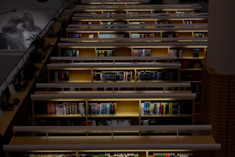 A library with rows of wooden bookshelves filled with a wide variety of books. Overhead lighting illuminates each shelf, creating a warm and inviting atmosphere. A few potted plants add a touch of greenery to the environment. A railing and staircase are visible on one side.