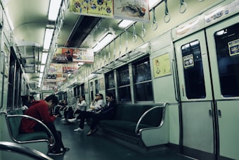 An interior of a subway train car with several passengers seated, some looking at their phones and others relaxing. The train is dimly lit with greenish hues dominating the seating and the walls. Advertisements hang from the ceiling, written in a language that appears to be Japanese.