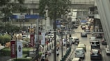 A busy urban scene featuring a crowded street with heavy traffic, including cars and taxis. Pedestrians are walking on the sidewalks, and various signs and advertisements are visible. A large overpass runs above the street with train tracks. Trees and urban greenery add some natural elements to the scene.