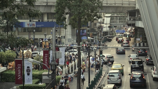 A busy urban scene featuring a crowded street with heavy traffic, including cars and taxis. Pedestrians are walking on the sidewalks, and various signs and advertisements are visible. A large overpass runs above the street with train tracks. Trees and urban greenery add some natural elements to the scene.