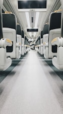 An empty train aisle with rows of modern, cushioned seats on either side. The interior is clean and well-lit with overhead reading lights, and there are display screens mounted above. The flooring is speckled, contributing to a sleek and practical design.