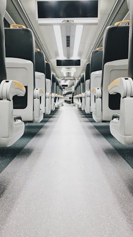 An empty train aisle with rows of modern, cushioned seats on either side. The interior is clean and well-lit with overhead reading lights, and there are display screens mounted above. The flooring is speckled, contributing to a sleek and practical design.