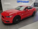 A vibrant red sports car is parked indoors on a tiled floor. The sleek vehicle has shiny black wheels and features a '5.0' badge on its side, indicating a powerful engine. The background includes signage related to vehicle technology and protection systems, with the words 'Opti-Coat' prominently displayed above the car.