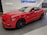 A vibrant red sports car is parked indoors on a tiled floor. The sleek vehicle has shiny black wheels and features a '5.0' badge on its side, indicating a powerful engine. The background includes signage related to vehicle technology and protection systems, with the words 'Opti-Coat' prominently displayed above the car.