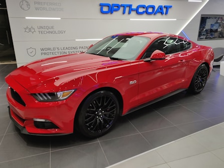 A vibrant red sports car is parked indoors on a tiled floor. The sleek vehicle has shiny black wheels and features a '5.0' badge on its side, indicating a powerful engine. The background includes signage related to vehicle technology and protection systems, with the words 'Opti-Coat' prominently displayed above the car.