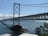 A large suspension bridge spans across a body of water, connecting two land masses. The bridge features a series of cables and towers, with a distinct structural design. Below the bridge, the water is a deep blue, with visible whitecaps and waves. In the foreground, green foliage and a rocky outcrop are visible.