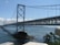 A large suspension bridge spans across a body of water, connecting two land masses. The bridge features a series of cables and towers, with a distinct structural design. Below the bridge, the water is a deep blue, with visible whitecaps and waves. In the foreground, green foliage and a rocky outcrop are visible.