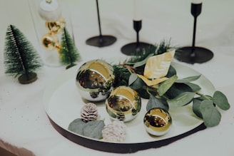 A decorative arrangement featuring shiny gold ornaments placed on a white tray, accompanied by green foliage and pine cones. Small artificial Christmas trees and black candle holders in the background enhance the festive setting.