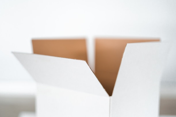 A close-up view of an open, empty cardboard box with a white exterior. The box flaps are unfolded, revealing the brown interior. The focus is on the box, with a minimalist, blurred white background.