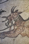 An ancient mosaic artwork depicting a mythical creature, possibly a dragon or griffin, holding a smaller animal in its mouth. The intricate tiles form a detailed and stylized representation with flowing lines and a sense of movement.