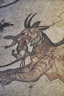 An ancient mosaic artwork depicting a mythical creature, possibly a dragon or griffin, holding a smaller animal in its mouth. The intricate tiles form a detailed and stylized representation with flowing lines and a sense of movement.