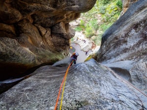 A person wearing a helmet is rappelling down a steep rock face in a canyon. The ropes are brightly colored, and there are several people visible below, indicating a group activity. The surroundings are rocky with patches of greenery visible.