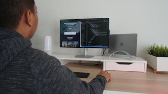 A person in a hoodie is sitting at a desk using a computer monitor. The screen displays coding software, and there is a closed laptop on a stand next to the monitor. The desk is organized, with a potted plant on the right and a small speaker on the left. The person is interacting with the computer using a wireless keyboard and mouse.