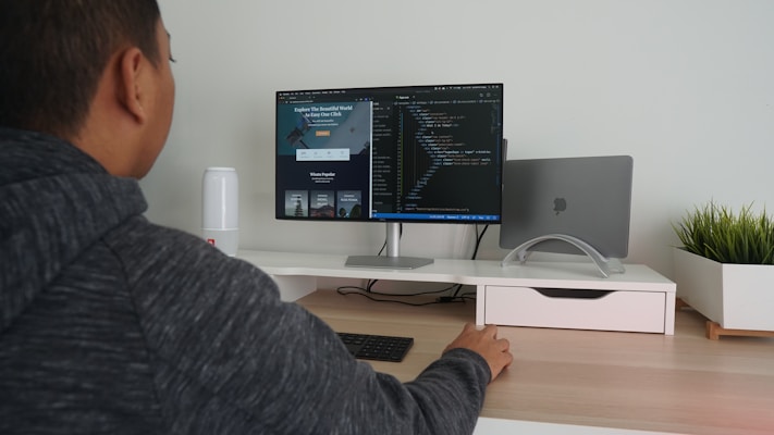 A person in a hoodie is sitting at a desk using a computer monitor. The screen displays coding software, and there is a closed laptop on a stand next to the monitor. The desk is organized, with a potted plant on the right and a small speaker on the left. The person is interacting with the computer using a wireless keyboard and mouse.