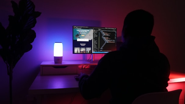 A person is sitting in a dark room, facing a computer screen that displays code. A desk lamp casts a blue glow, contrasting with the reddish ambient lighting. A silhouette of the person is visible, and there is a plant partially seen on the left.