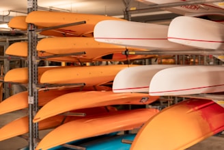 Several orange and white kayaks are stored on metal racks inside a storage area. The kayaks are neatly stacked in rows, with the orange kayaks predominantly on the left side and the white ones on the right. The scene is indoors, and the lighting is warm, creating a cozy atmosphere. Metal beams and supports are visible, securely holding the kayaks in place.