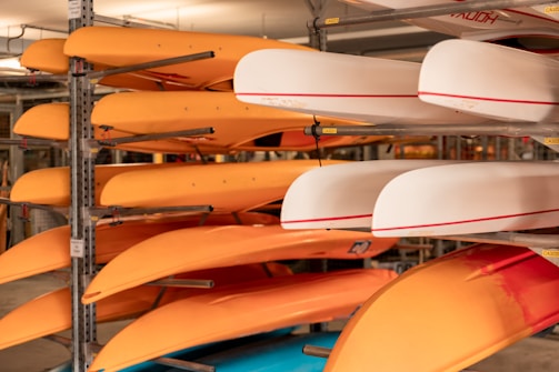 Several orange and white kayaks are stored on metal racks inside a storage area. The kayaks are neatly stacked in rows, with the orange kayaks predominantly on the left side and the white ones on the right. The scene is indoors, and the lighting is warm, creating a cozy atmosphere. Metal beams and supports are visible, securely holding the kayaks in place.