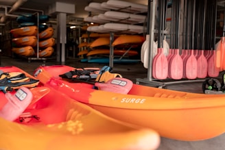 A collection of bright orange kayaks with black straps are stacked in a storage area. Paddles with pink blades are neatly organized on the right side. In the background, additional kayaks are stored on racks, creating a vibrant display. The setting appears to be an indoor facility, with paddleboards and various accessories visible.