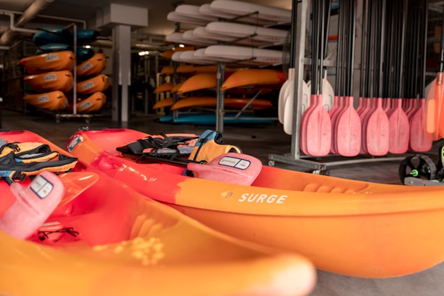 A collection of bright orange kayaks with black straps are stacked in a storage area. Paddles with pink blades are neatly organized on the right side. In the background, additional kayaks are stored on racks, creating a vibrant display. The setting appears to be an indoor facility, with paddleboards and various accessories visible.