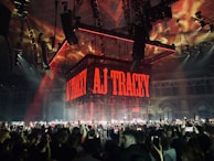 A large indoor concert venue is filled with a crowd of people holding up smartphones, capturing the performance. A prominent stage setup includes a suspended cube-shaped digital screen displaying the name AJ Tracey in bold, red letters. The ceiling and structures are illuminated with dynamic red and orange lighting.