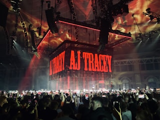A large indoor concert venue is filled with a crowd of people holding up smartphones, capturing the performance. A prominent stage setup includes a suspended cube-shaped digital screen displaying the name AJ Tracey in bold, red letters. The ceiling and structures are illuminated with dynamic red and orange lighting.