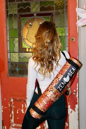A person with long, wavy hair stands facing a detailed stained glass and worn wooden door, carrying a colorful yoga mat with a floral design on their back. The person is wearing a white long-sleeve top and dark pants.