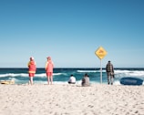A beach scene featuring two lifeguards in yellow and red uniforms standing on the sand, facing the ocean. A sign warns of a dangerous current. Nearby, a small group of people are seated, observing the waves. A lifeguard boat is visible on the sand.