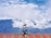 A rooftop with a red tiled design featuring a decorative wind vane shaped like a horse and rider is seen against a backdrop of lush green mountains and a partly cloudy blue sky.