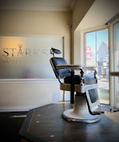An empty barber chair with a vintage design is positioned on a wooden platform inside a barber shop. The chair features black upholstery and metallic armrests. A frosted glass window with signage for 'Starks Barber Company' is situated behind the chair, and large windows allow natural light to illuminate the space, offering a view of the street outside.